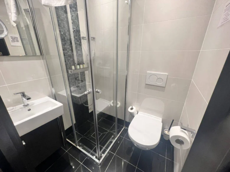 Modern bathroom with black tile floor, white walls, a glass shower enclosure, wall-mounted toilet with dual flush buttons, toilet paper holder, and a white sink with a mirror above it. Toiletries are visible in the shower.