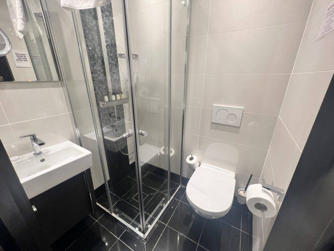 Modern bathroom with black tile floor, white walls, a glass shower enclosure, wall-mounted toilet with dual flush buttons, toilet paper holder, and a white sink with a mirror above it. Toiletries are visible in the shower.
