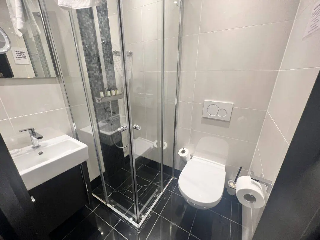 Modern bathroom with black tile floor, white walls, a glass shower enclosure, wall-mounted toilet with dual flush buttons, toilet paper holder, and a white sink with a mirror above it. Toiletries are visible in the shower.