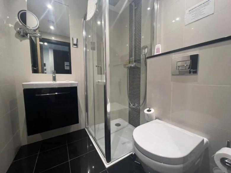 Modern bathroom with white walls, dark floor tiles, a shower with clear glass doors, a wall-mounted sink and mirror on the left, and a toilet with a toilet paper roll on the right.