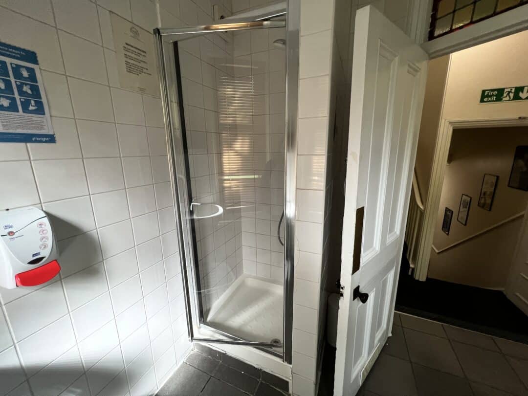 A small shower cubicle with a glass door is located in a tiled bathroom. A white door is open next to it, and a wall-mounted soap dispenser is visible on the left. Stairs and framed pictures are seen through the doorway.