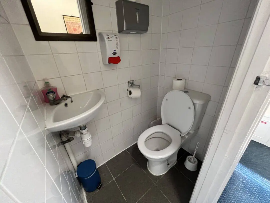 A small restroom with a white toilet, toilet brush, wall-mounted sink, soap dispenser, mirror, paper towel dispenser, and a blue trash bin on a dark tiled floor with white tiled walls.