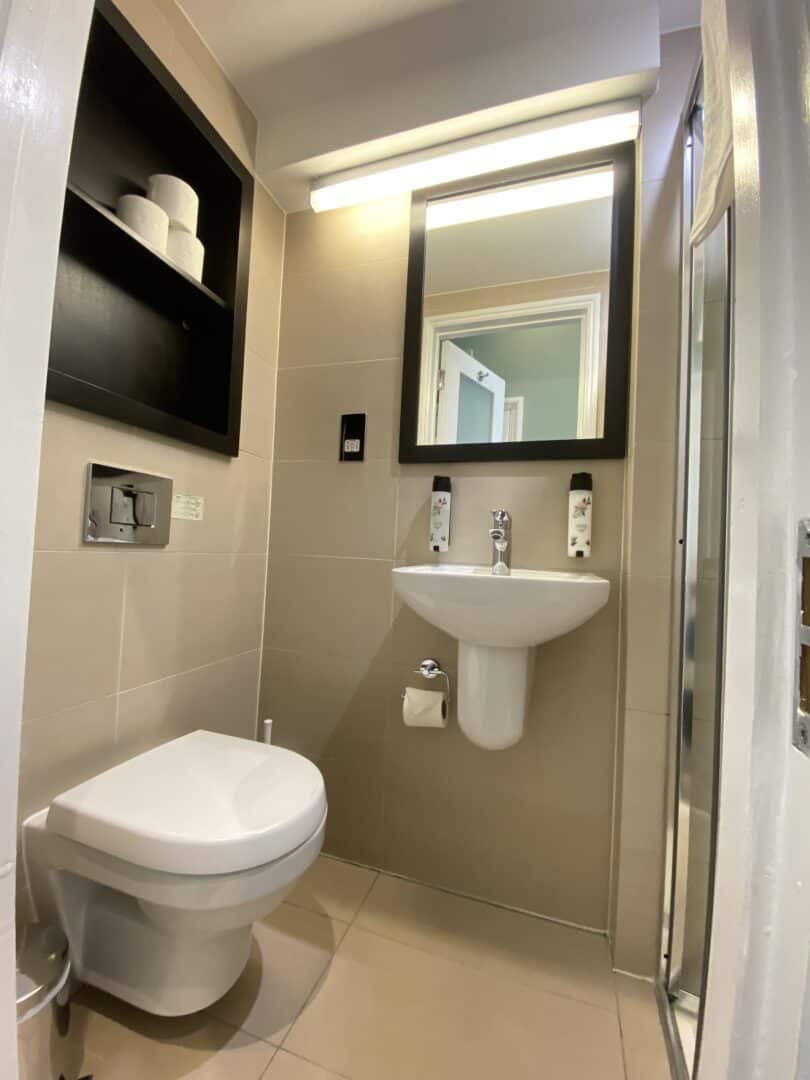 A modern compact bathroom with a wall-mounted toilet, floating sink, mirror, shelf with toilet paper, soap dispensers above the sink, and a glass shower door visible on the right.