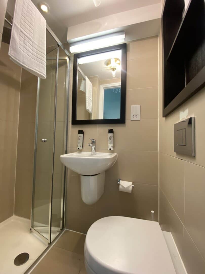 A modern, compact bathroom with a glass shower, wall-mounted sink, toilet, mirror, towel rack, and beige tiles. The lighting is bright and shelving is visible on the right wall.