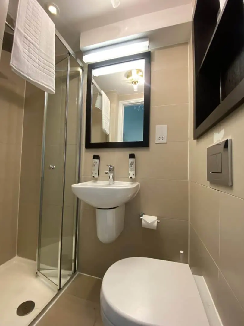 A modern, compact bathroom with a glass shower, wall-mounted sink, toilet, mirror, towel rack, and beige tiles. The lighting is bright and shelving is visible on the right wall.