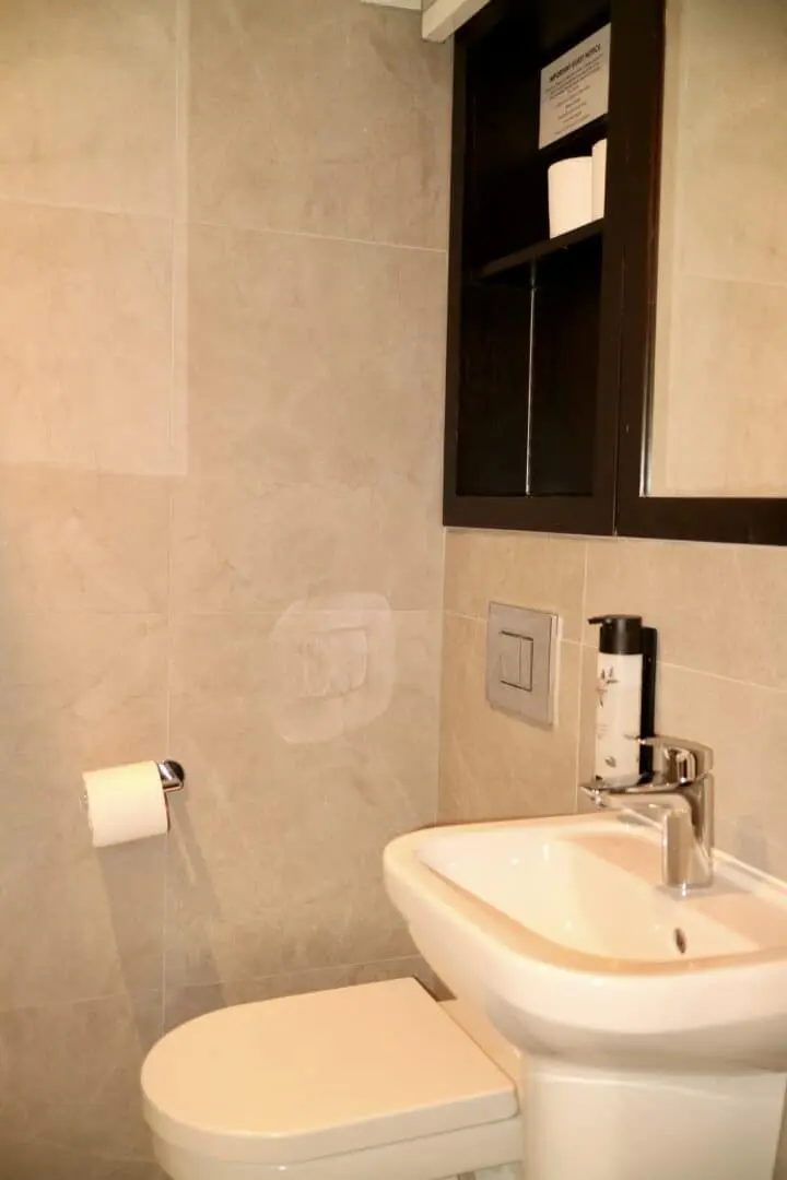 A modern bathroom with light gray tiled walls, featuring a white toilet and sink. A toilet paper roll is mounted on the wall, and a shelf above holds extra toilet paper and a framed sign.