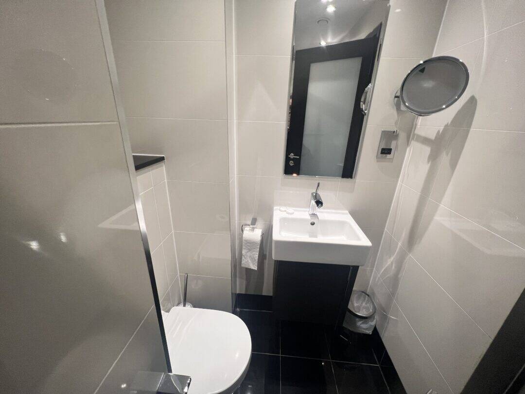 A modern bathroom with a toilet, a wall-mounted sink, a rectangular mirror above the sink, a round magnifying mirror, a towel on a rack, a small bin, and glossy black floor tiles.