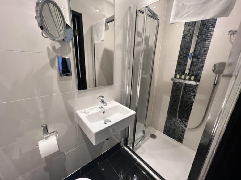 Modern bathroom with white tiled walls, a small wall-mounted sink, mirror, toilet paper holder, and a glass shower enclosure with black mosaic tiles and toiletries on a shelf.