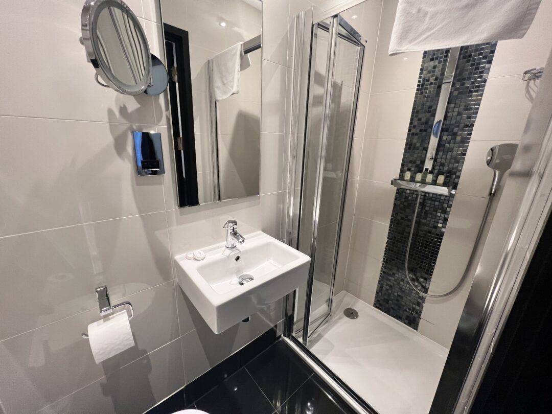 Modern bathroom with white tiled walls, a small wall-mounted sink, mirror, toilet paper holder, and a glass shower enclosure with black mosaic tiles and toiletries on a shelf.