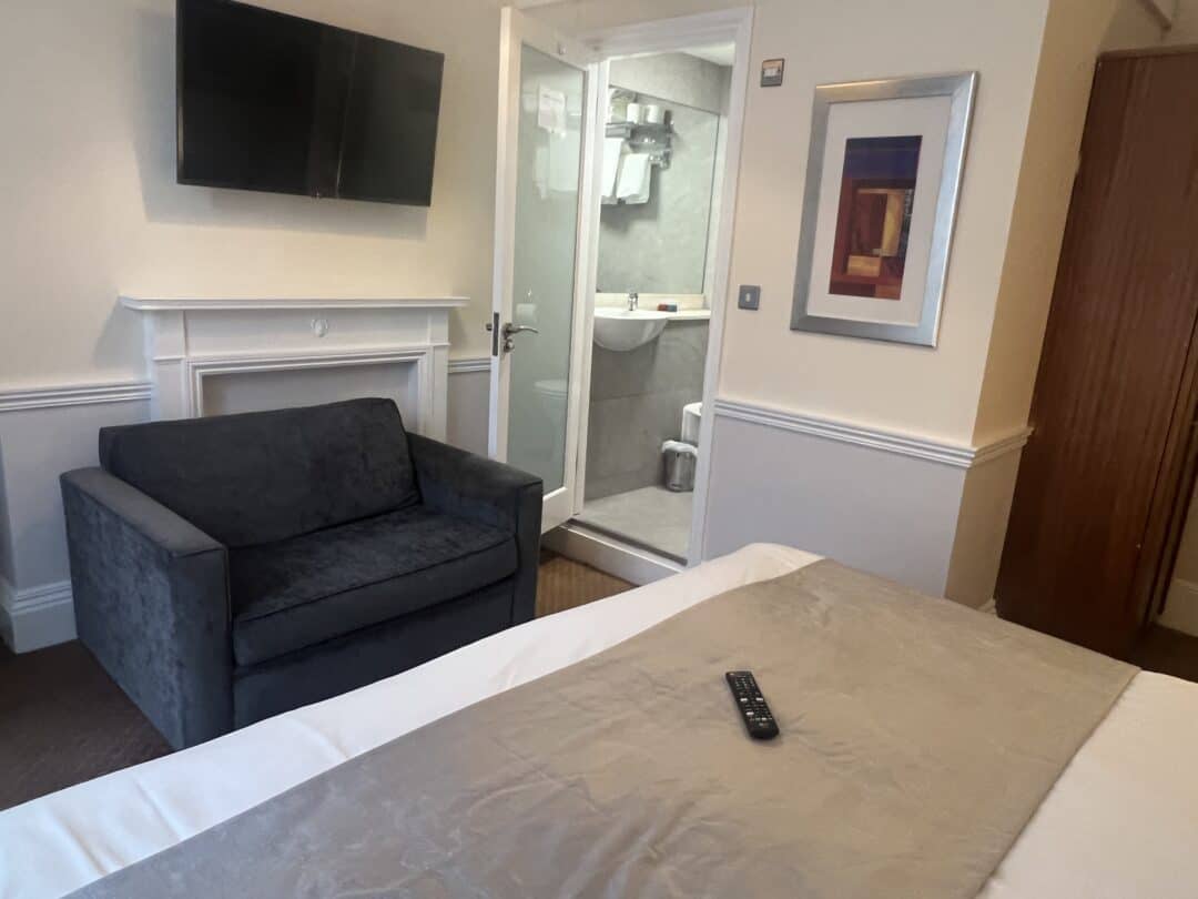 A hotel room with a bed, a gray loveseat, a wall-mounted TV, a framed artwork, and an open door leading to a small bathroom with a sink and mirror. A remote control rests on the bed.