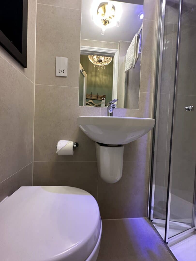 A small bathroom with beige tiles, a wall-mounted sink and toilet, a glass shower enclosure, a toilet paper holder, and a mirror reflecting a chandelier and coat rack.