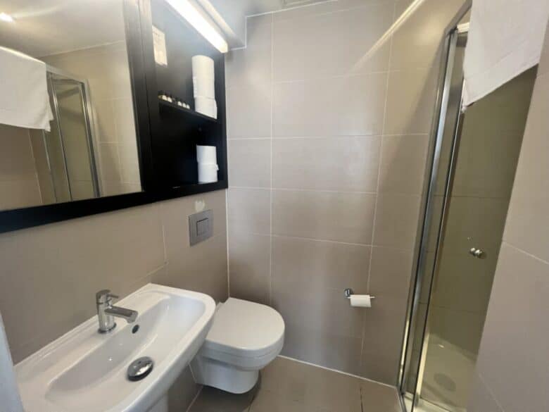 A small modern bathroom with beige tiles, a wall-mounted sink, a toilet, a mirror cabinet with toiletries and towels, a toilet paper holder, and a glass shower enclosure.