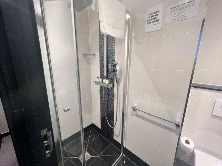 A modern glass shower with black and white tiles, a rainfall showerhead, mounted toiletries, a white towel, and a notice on the wall. A towel rack and toilet paper are visible outside the shower.
