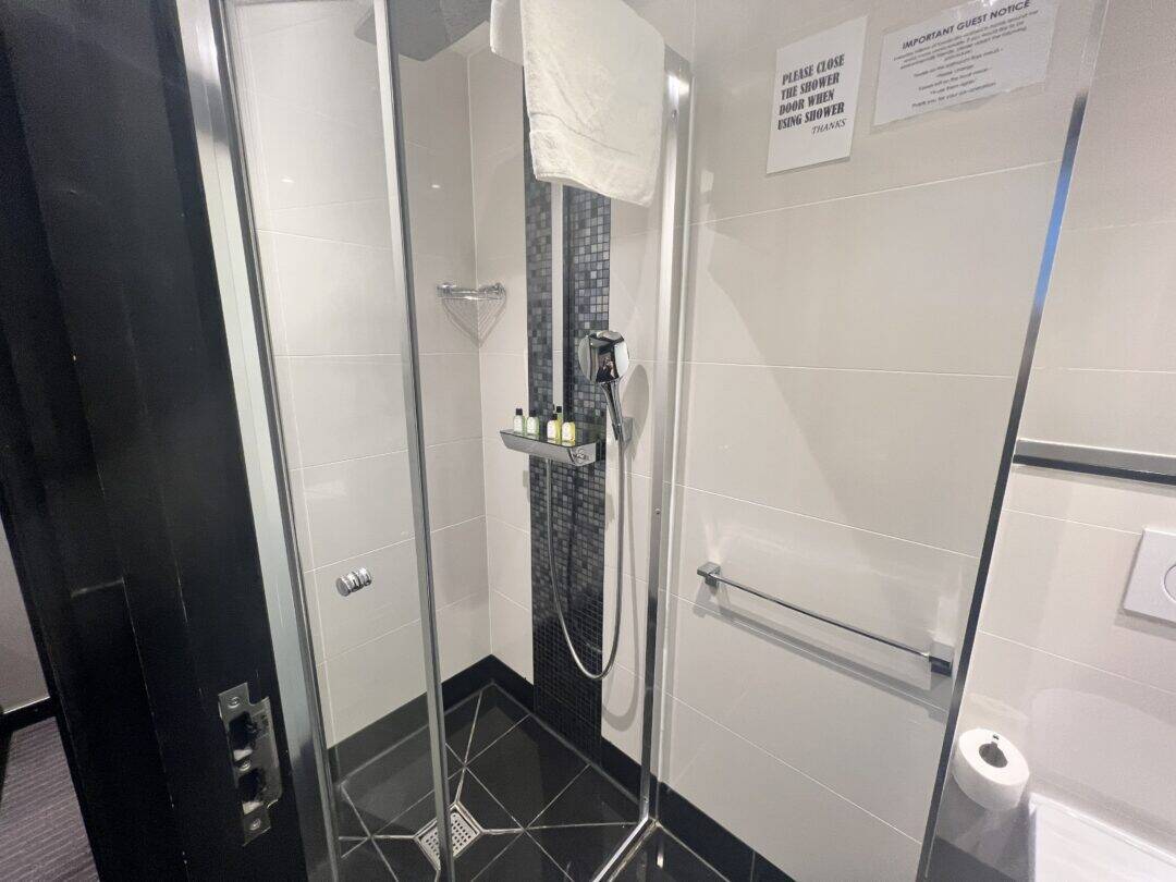 A modern glass shower with black and white tiles, a rainfall showerhead, mounted toiletries, a white towel, and a notice on the wall. A towel rack and toilet paper are visible outside the shower.