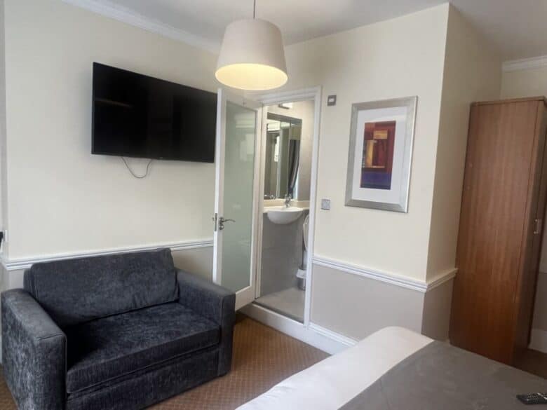 A small hotel room with a gray sofa, wall-mounted TV, wardrobe, bed, and a compact bathroom visible through an open door. Modern art hangs on a cream-colored wall beneath a ceiling lamp.