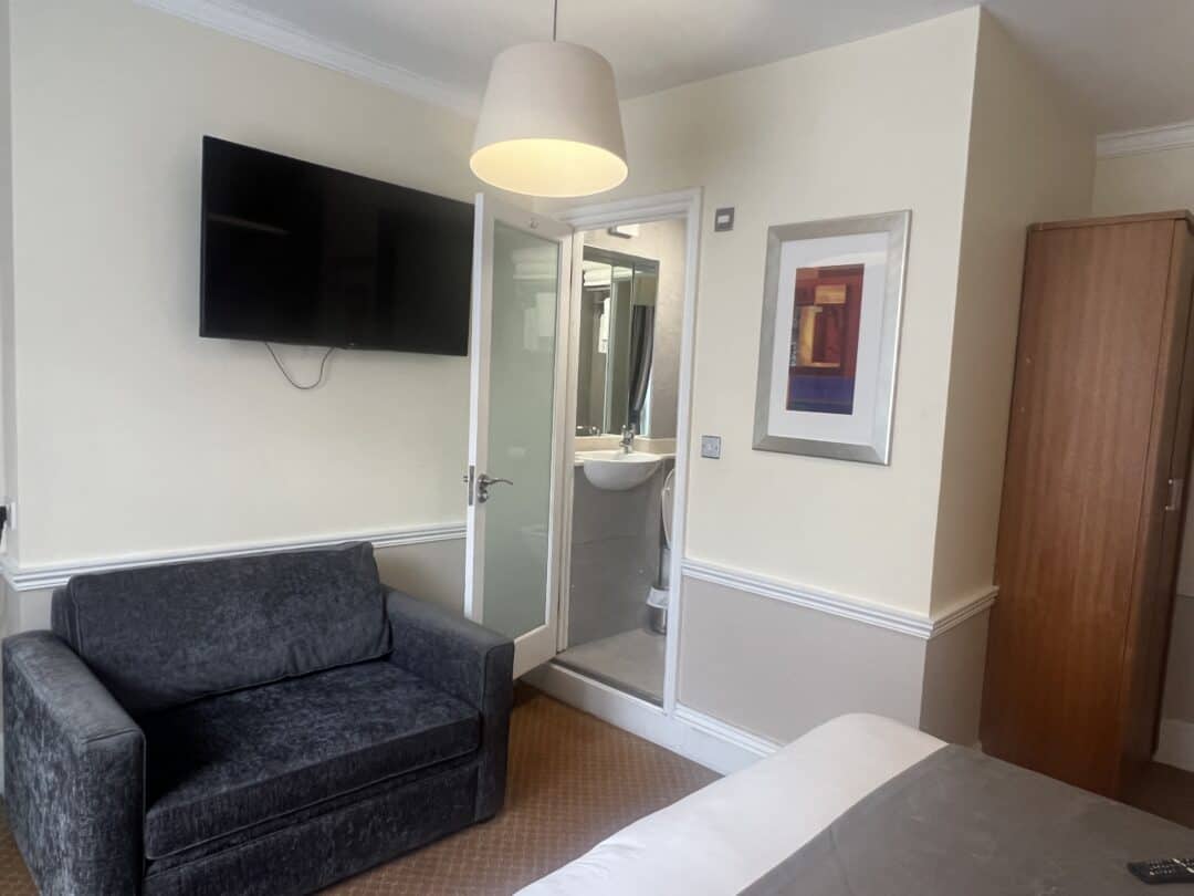 A small hotel room with a gray sofa, wall-mounted TV, wardrobe, bed, and a compact bathroom visible through an open door. Modern art hangs on a cream-colored wall beneath a ceiling lamp.