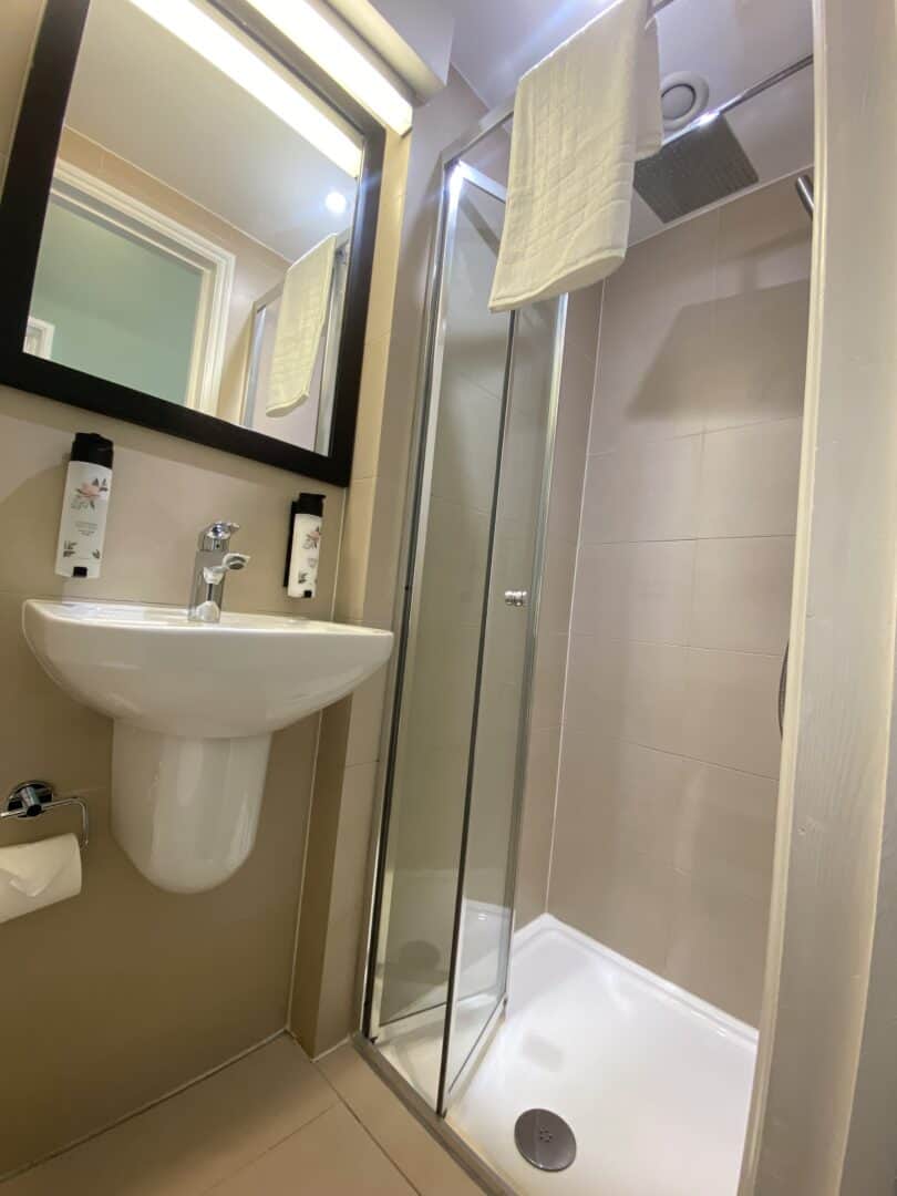 A compact bathroom with a wall-mounted sink, mirror, glass shower enclosure, overhead shower, towel hanging above, and soap dispensers on the wall next to the sink.
