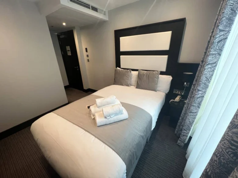 A neatly made bed with gray and white bedding and three folded towels on top sits in a modern hotel room with neutral walls, a bedside lamp, and a window with patterned curtains.