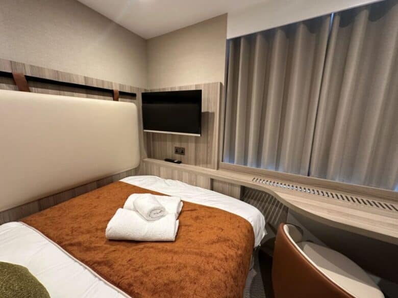 A small, modern hotel room with a double bed, brown blanket, two folded white towels, a wall-mounted TV, and closed curtains over a window. A desk and chair are positioned beside the bed.