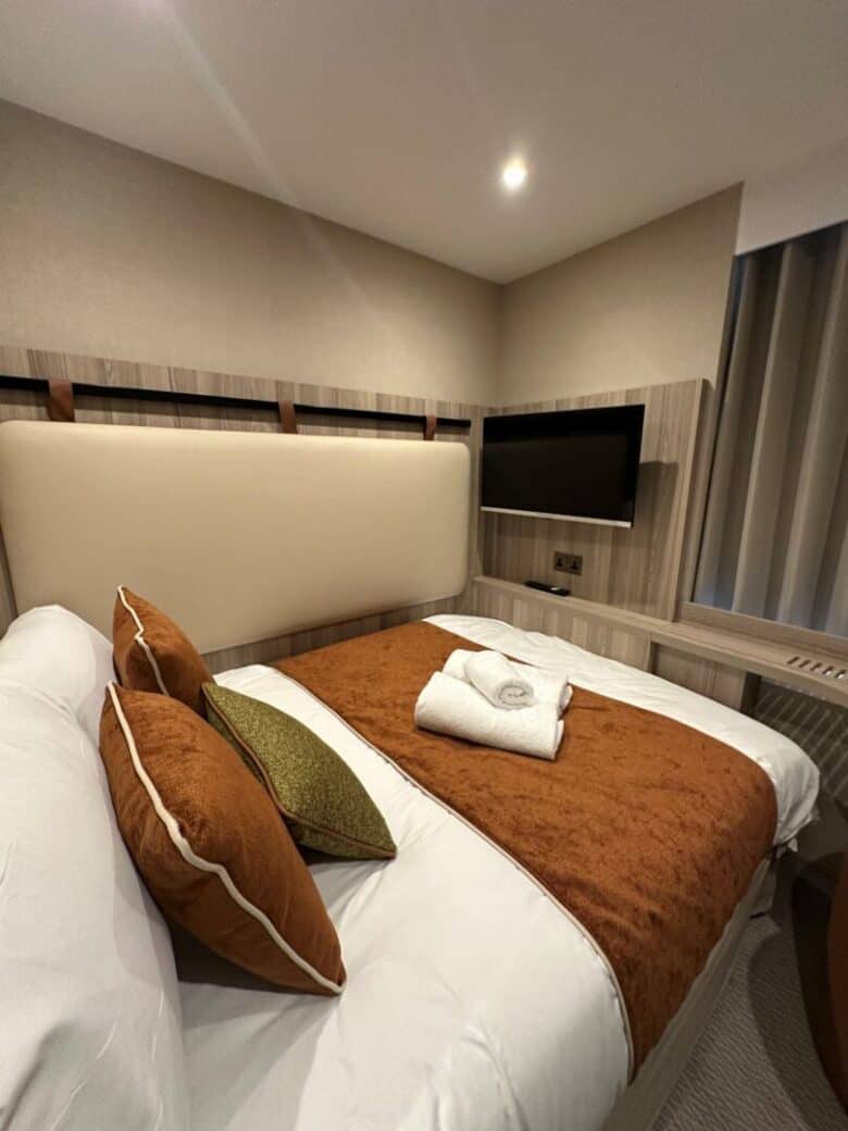 A neatly made bed with brown and green accent pillows and a folded towel sits in a modern hotel room with neutral tones, a mounted flat-screen TV, and soft lighting.