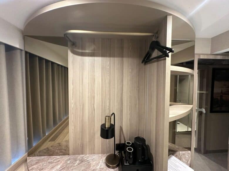 A small modern hotel room closet with two black hangers, a clothes rail, a lamp, and a coffee machine on a marble countertop below, with mirrors and vertical blinds reflected on the left.