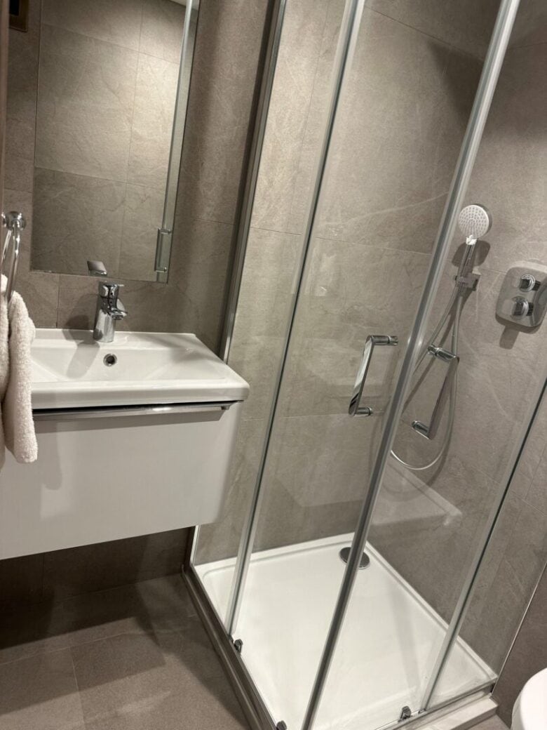 A modern bathroom with beige tiles, featuring a white sink with a faucet, a wall-mounted towel, and a glass shower enclosure with a handheld showerhead and soap dispenser.