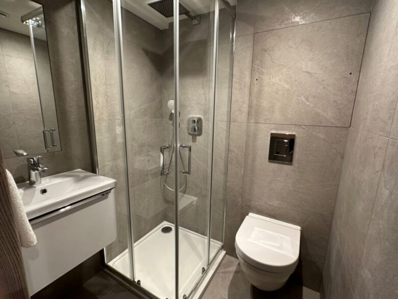 Modern bathroom with gray tiled walls, a glass-enclosed corner shower, a white wall-mounted sink with a towel, and a wall-mounted toilet. The space appears compact and clean.