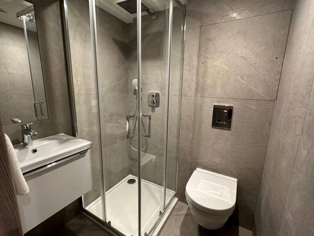 Modern bathroom with gray tiled walls, a glass-enclosed corner shower, a white wall-mounted sink with a towel, and a wall-mounted toilet. The space appears compact and clean.