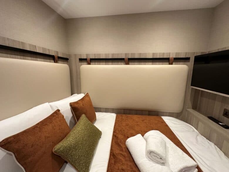 A modern, compact bedroom with a double bed, brown and green accent pillows, a brown throw, and two neatly folded white towels. Light wood paneling, beige walls, and a mounted flat-screen TV are visible.