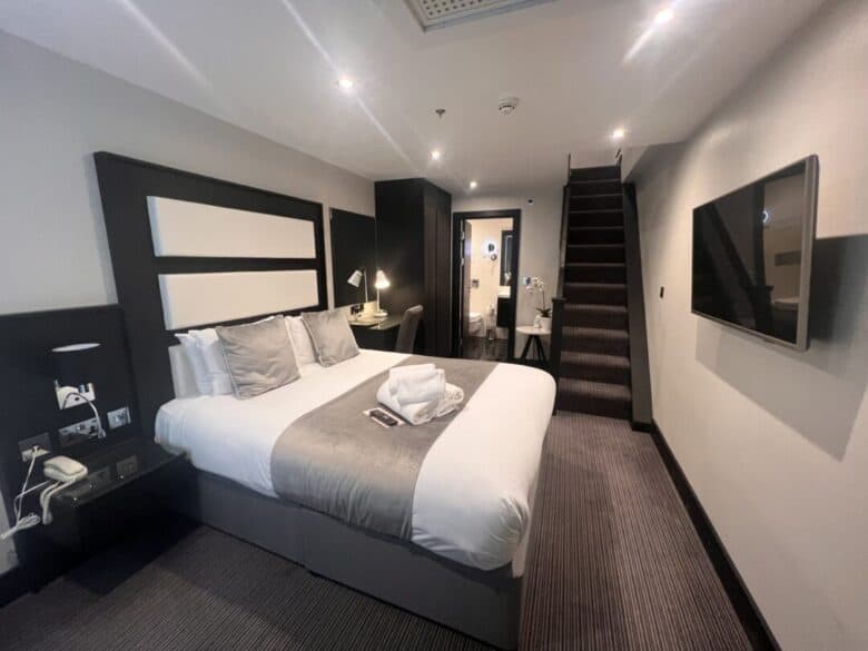 Modern hotel room with a large double bed, gray and white decor, a desk with a chair, wall-mounted TV, and stairs leading to an upper level. Towels and toiletries are neatly placed on the bed.