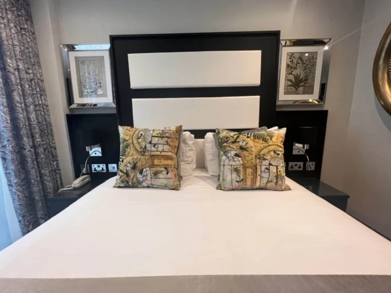 A neatly made bed with two decorative pillows featuring botanical prints, set against a modern black and white headboard, with bedside tables, framed artwork, and patterned curtains visible.