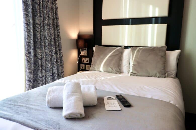 A neatly made hotel bed with gray and white bedding, three pillows, two rolled white towels, a TV remote, and a "please make up my room" door hanger on top. A window with patterned curtains and a bedside lamp are visible.