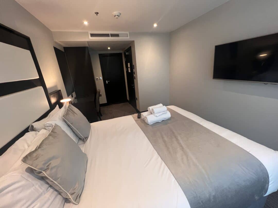 A modern hotel room with a neatly made double bed, gray and white bedding, two stacked towel sets on top, a wall-mounted TV, soft lighting, and a view of the hallway and dark entrance door.