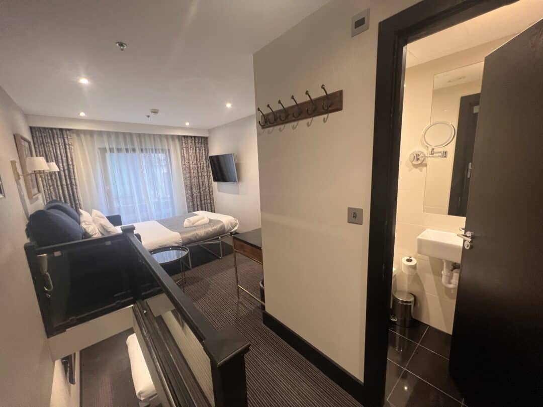 A modern hotel room with a bed, sofa, wall-mounted TV, and large window with curtains. An open door reveals a small bathroom with a sink and mirror. A coat rack is mounted on the wall near the entrance.