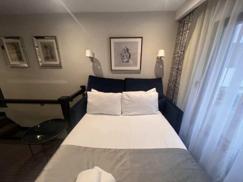 A neatly made bed with white pillows and a gray throw sits against a wall, flanked by wall-mounted lamps. Framed artwork hangs above the bed, and sheer curtains let in natural light from a window on the right.