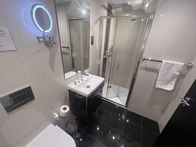 Modern bathroom with glossy black floor tiles, a glass shower enclosure, a white sink with a mirror above it, a mounted lit vanity mirror, a toilet, a towel on a rail, and a toilet paper roll.