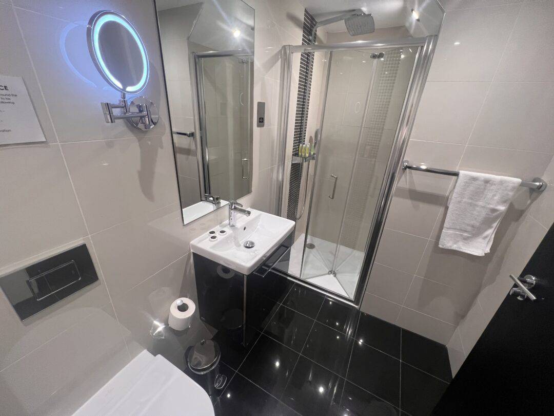 Modern bathroom with glossy black floor tiles, a glass shower enclosure, a white sink with a mirror above it, a mounted lit vanity mirror, a toilet, a towel on a rail, and a toilet paper roll.
