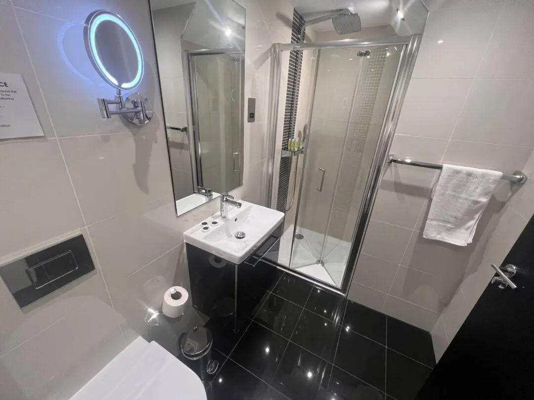 Modern bathroom with glossy black floor tiles, a glass shower enclosure, a white sink with a mirror above it, a mounted lit vanity mirror, a toilet, a towel on a rail, and a toilet paper roll.