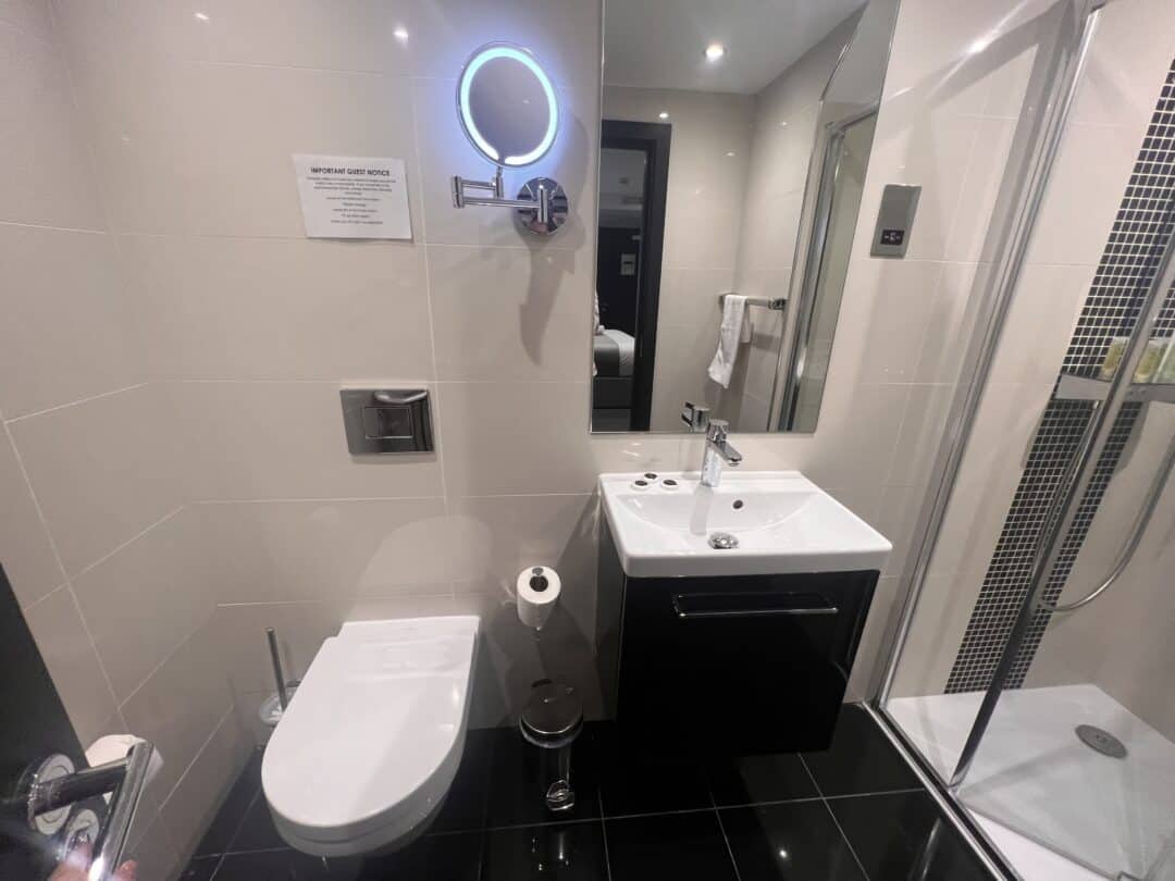 Modern bathroom with white walls, a wall-mounted toilet, a sink with a black cabinet, round lit mirror, towel rack, glass shower enclosure, and a sign above the toilet. The floor is shiny black tile.