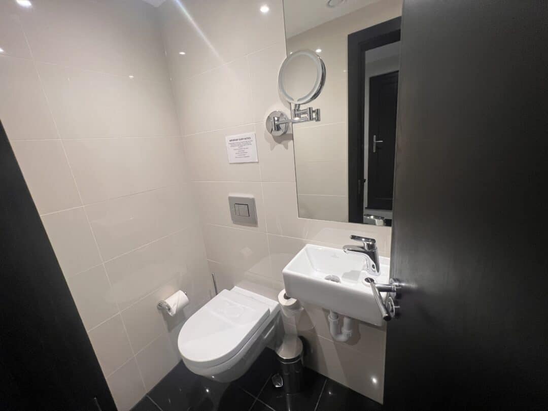 A small, modern bathroom with beige tiled walls, a wall-mounted toilet, a toilet paper holder, a small white sink, a round mirror mounted on the wall, and a partially open black door.