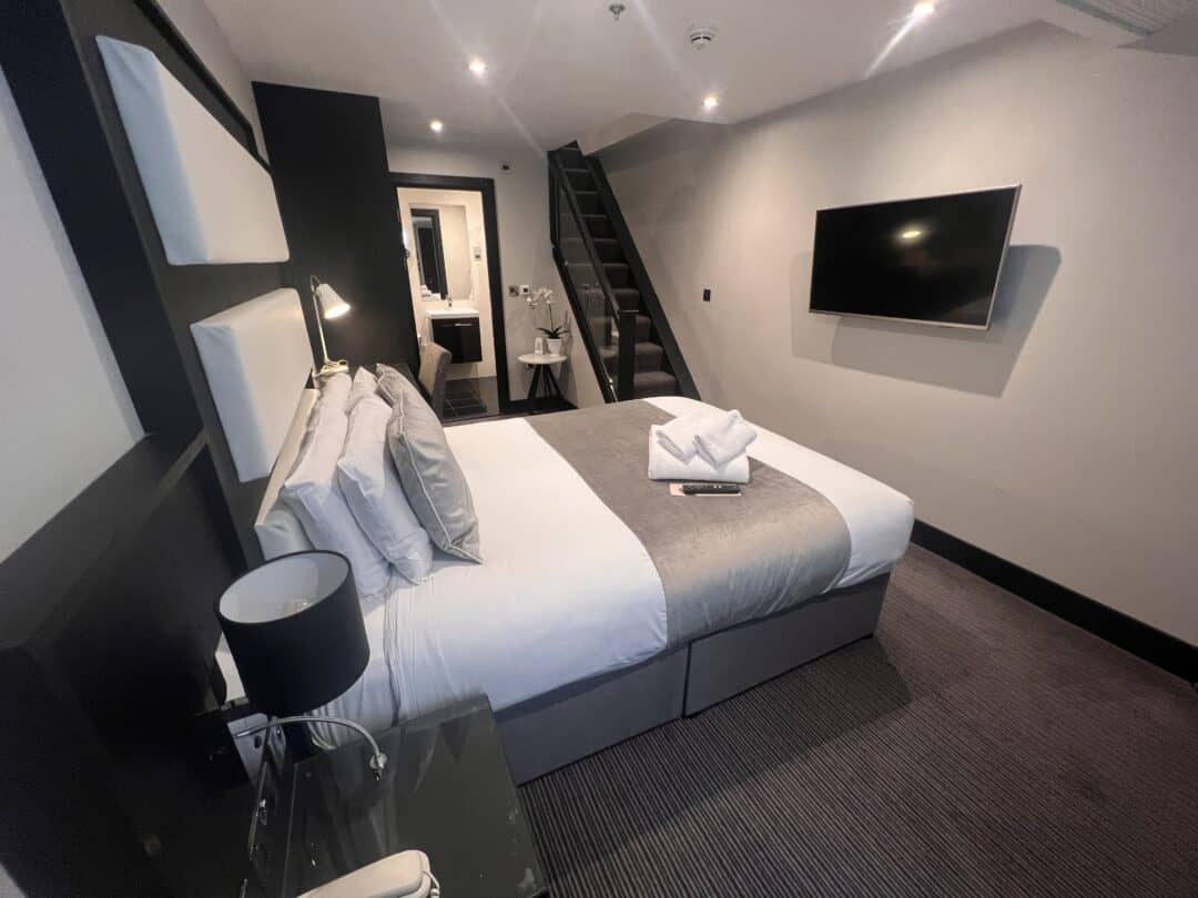 Modern hotel room with a double bed, gray and white bedding, folded towels, a wall-mounted TV, bedside table with a lamp, and a staircase leading to an upper level. The room has minimalist decor and muted colors.