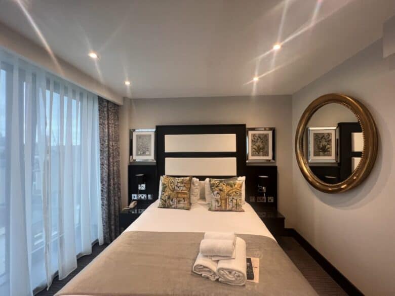 Modern hotel room with a neatly made double bed, decorative pillows, folded towels at the foot, large window with sheer curtains, round mirror on the wall, and framed artwork on either side of the bed.