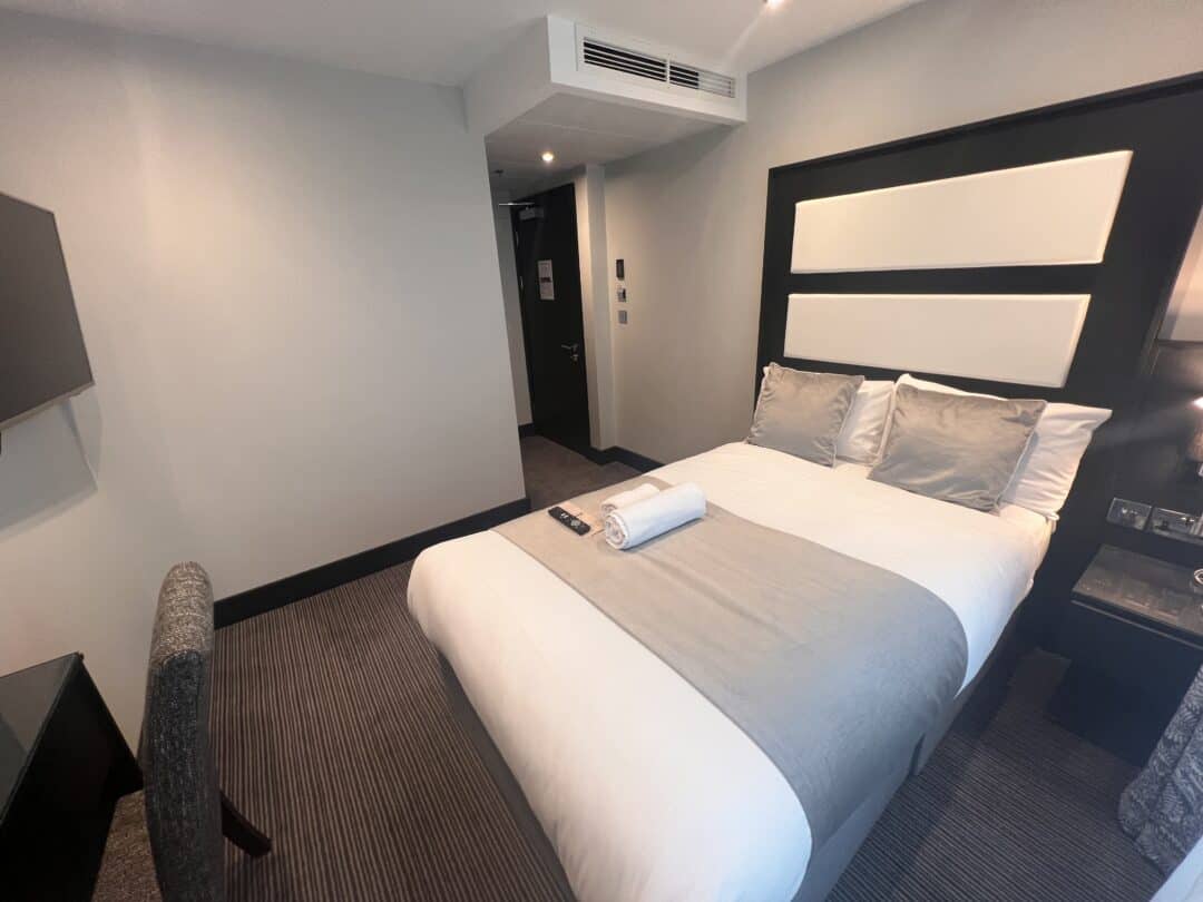 A modern hotel room with a neatly made double bed, gray and white bedding, two pillows, and a towel with a remote placed on top. There is a desk with a chair and a wall-mounted TV. The room has neutral colors and soft lighting.