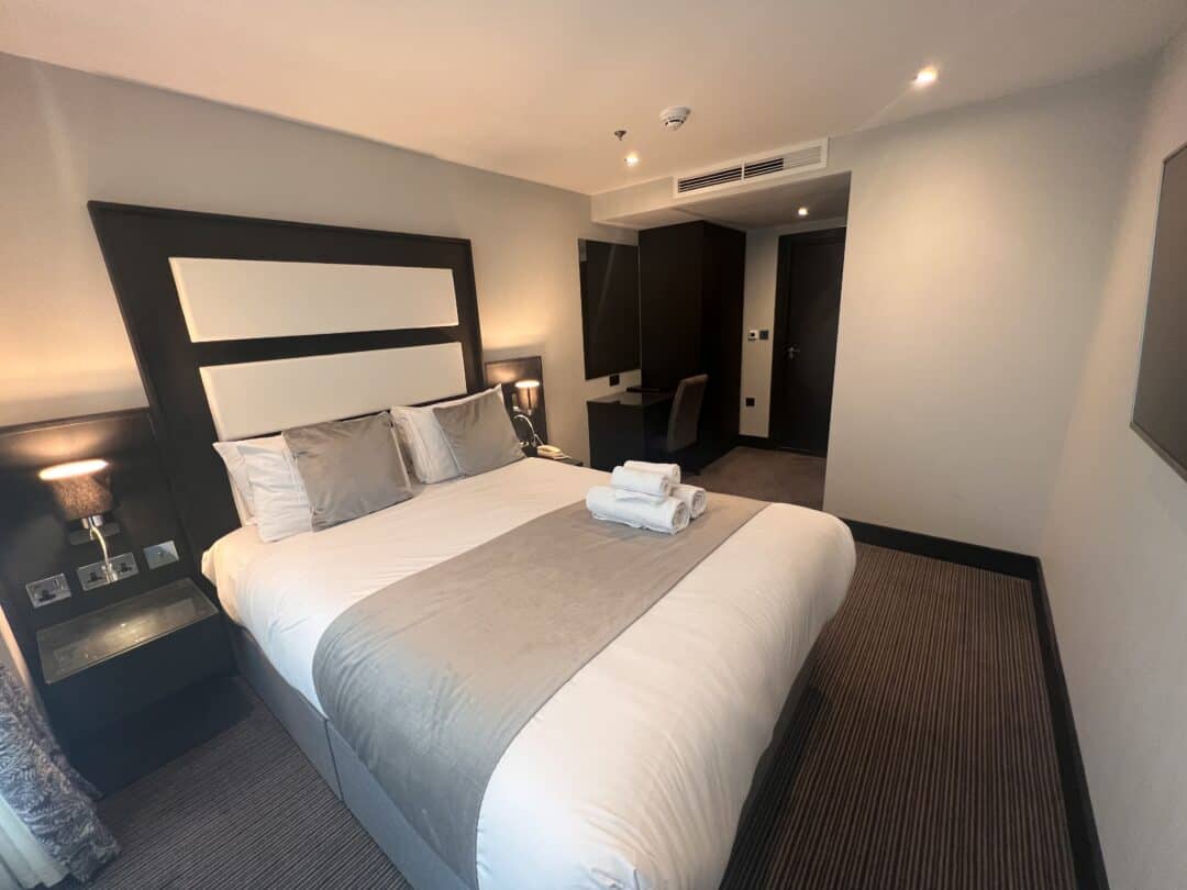 A modern hotel room with a double bed topped with gray and white bedding and neatly folded towels, two bedside tables with lamps, a desk and chair in the corner, and neutral decor with carpeted flooring.