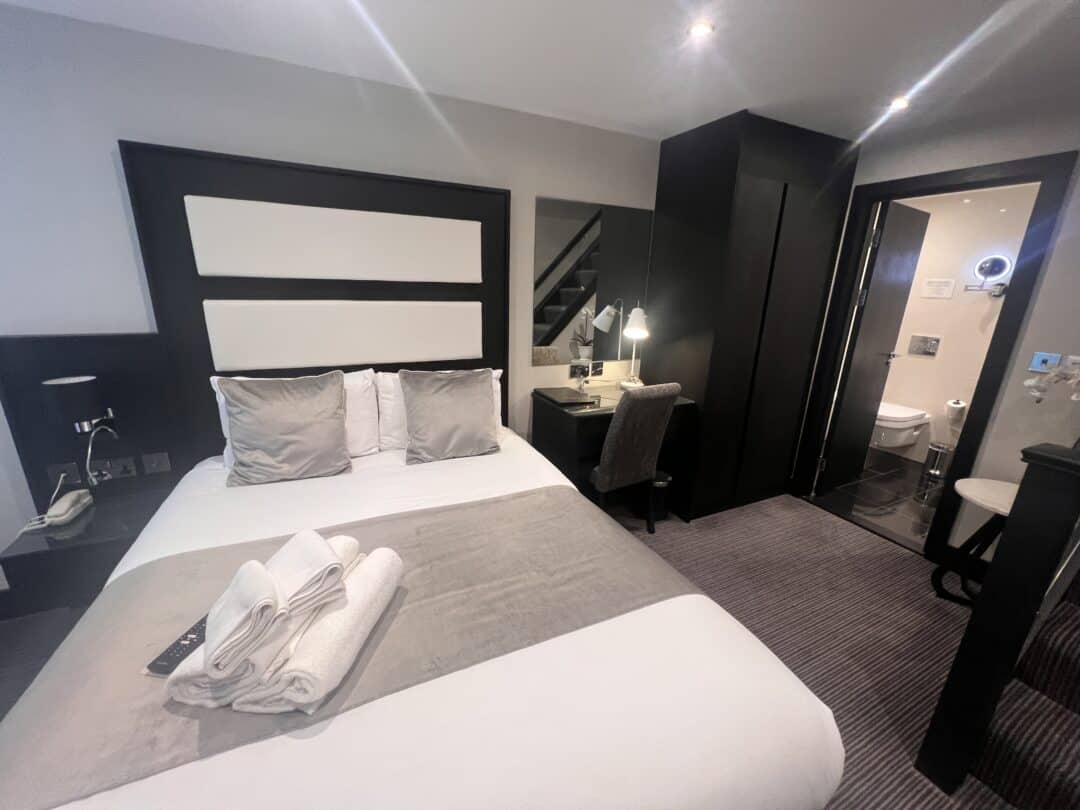 A modern hotel room with a double bed, neatly folded towels, a desk with a chair, a large mirror, and an en-suite bathroom with a toilet visible through an open door. The decor features neutral tones.
