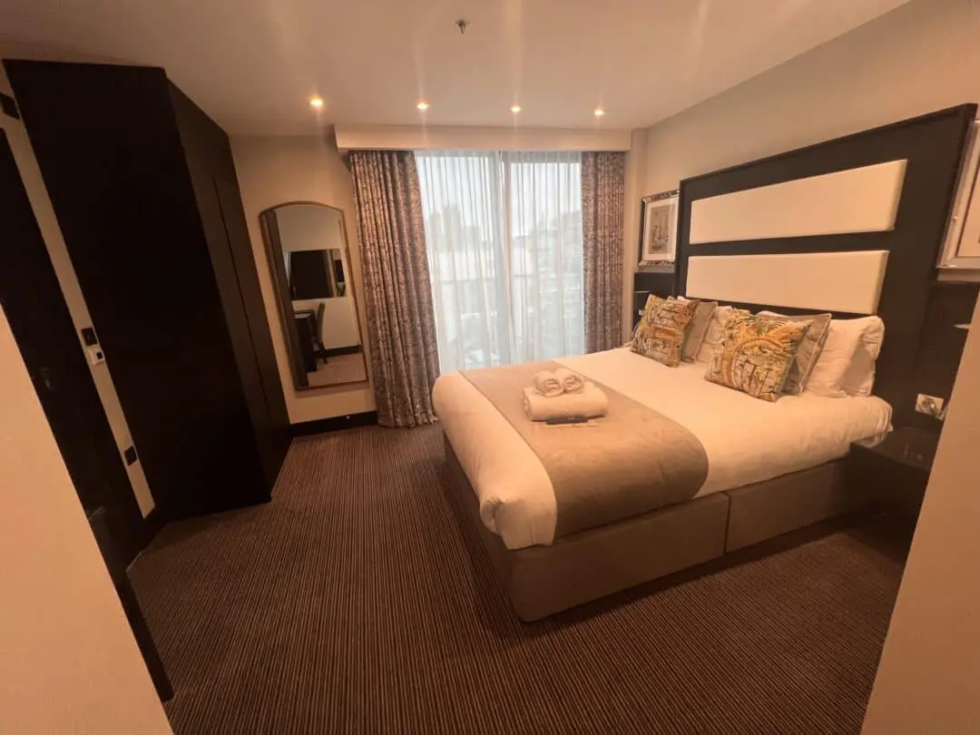 A neatly made double bed with decorative pillows and folded towels sits in a cozy hotel room with warm lighting, carpeted floor, a wardrobe, wall mirror, and large window with patterned curtains.
