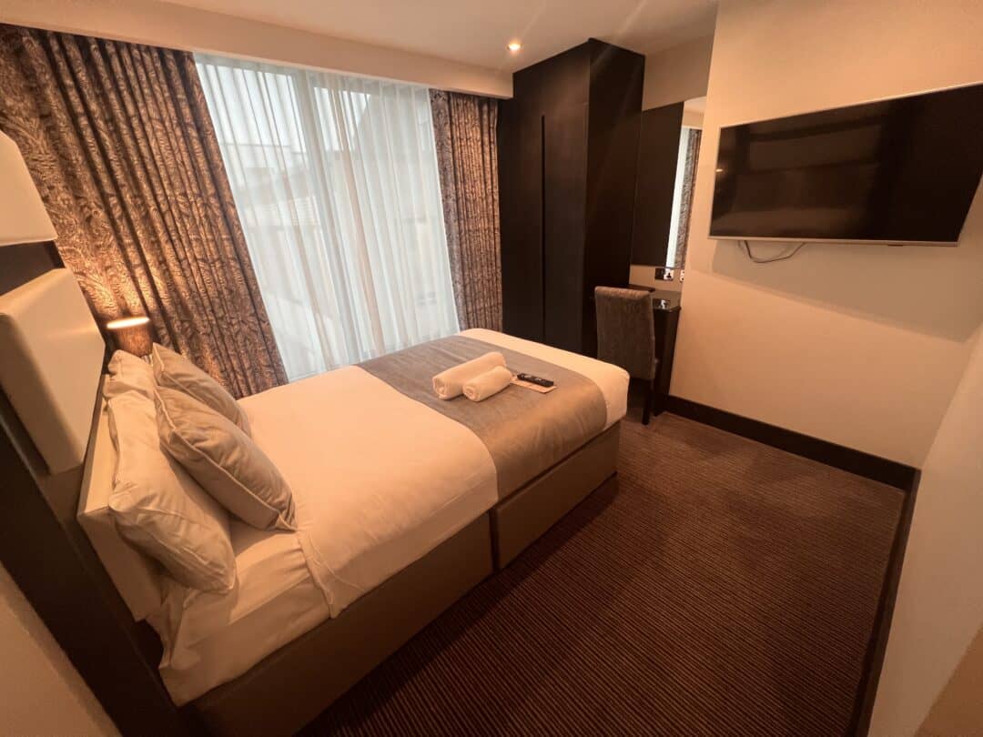 A neatly made single bed in a modern hotel room with brown carpet, large window with sheer curtains, mounted flat-screen TV, desk, and a chair. Towels and a remote control rest on the bed.