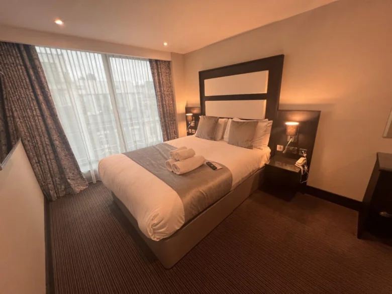 A neatly made double bed in a hotel room with two folded towels on top, flanked by nightstands and lamps, and a large window with sheer curtains letting in natural light.