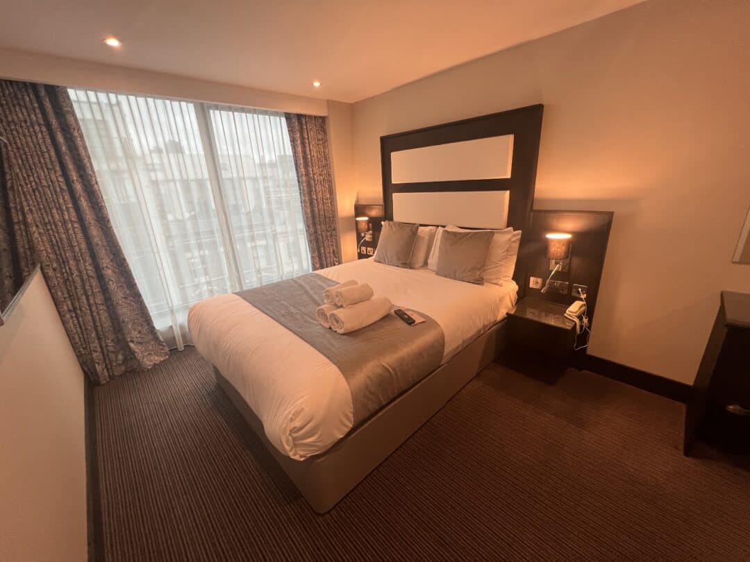 A neatly made double bed in a hotel room with two folded towels on top, flanked by nightstands and lamps, and a large window with sheer curtains letting in natural light.