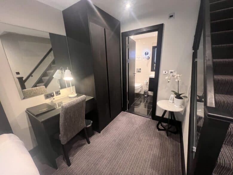 A modern hotel room with a desk and chair, a wall mirror, a wardrobe, a potted orchid on a small round table, carpeted stairs, and a view into a brightly lit bathroom.